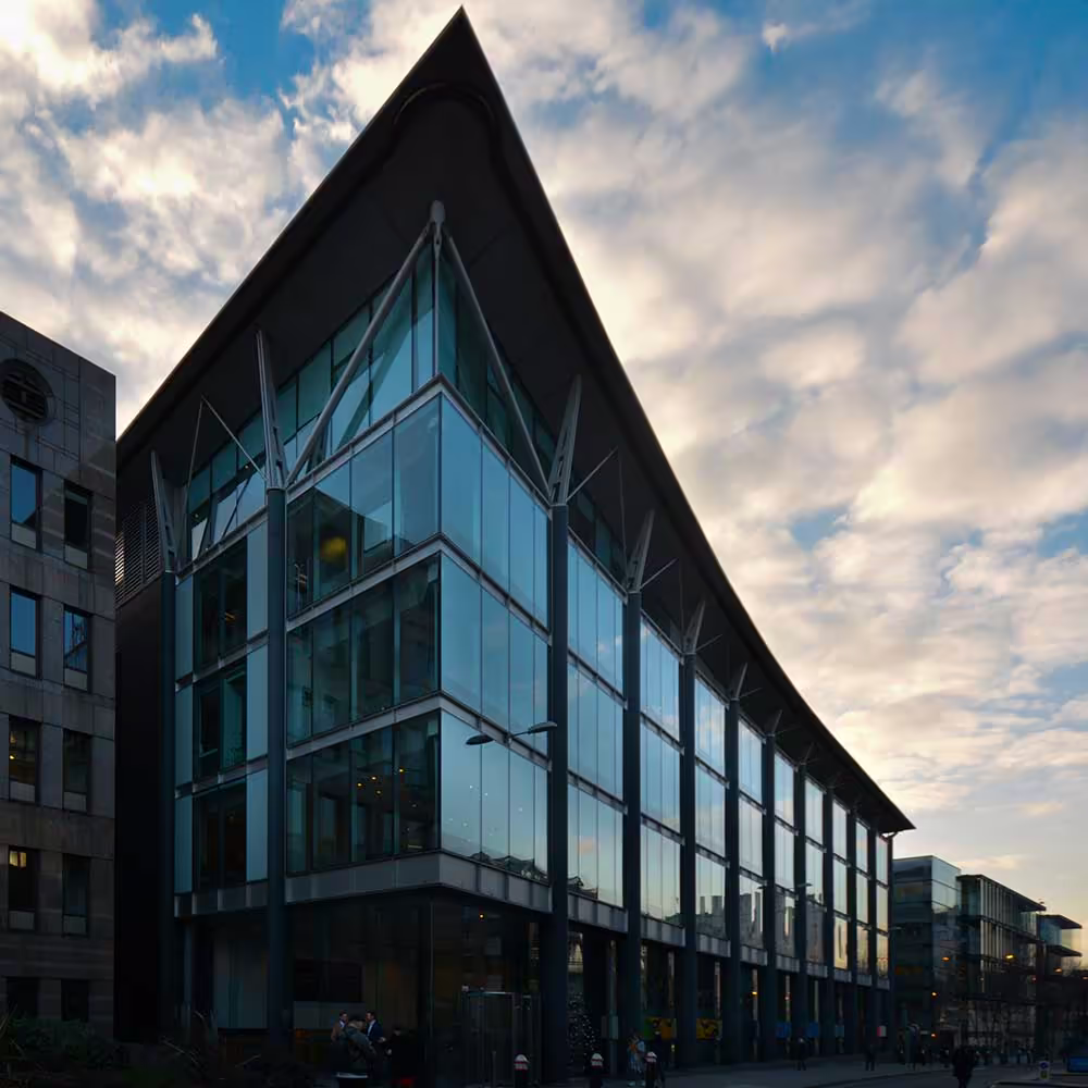 A wide, low-angle shot of the glass and steel modern office building with its distinctive sharp corner and curved roof, set against a dramatic sky with clouds, highlighting the commercial property's architecture.