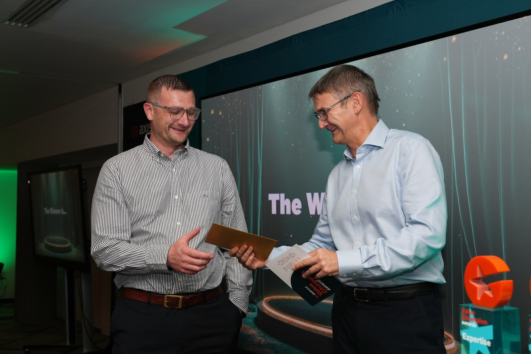 Man giving an award to someone. Handing a golden envelope. Two men. Trophy on the side