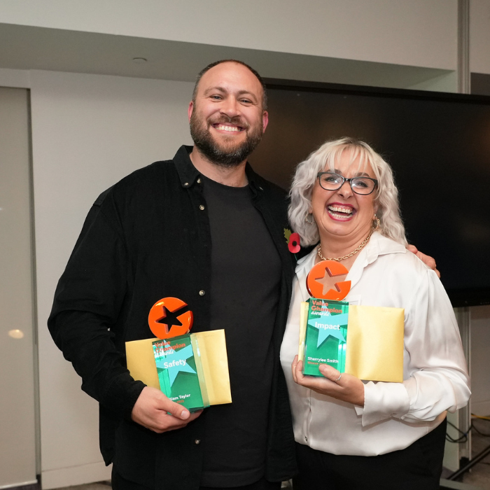 Man wearing dark shirt and t-shirt. Woman wearing white top. Both smiling and holding a gold envelope and a small rectangle blue and orange star trophy. Background in a grey room