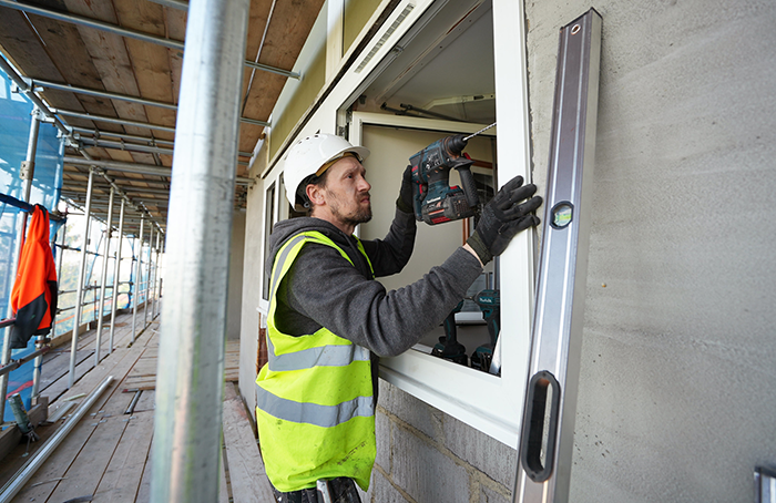 operative in high vis and hard hat using a drill to install window frame from exterior building scaffolding.