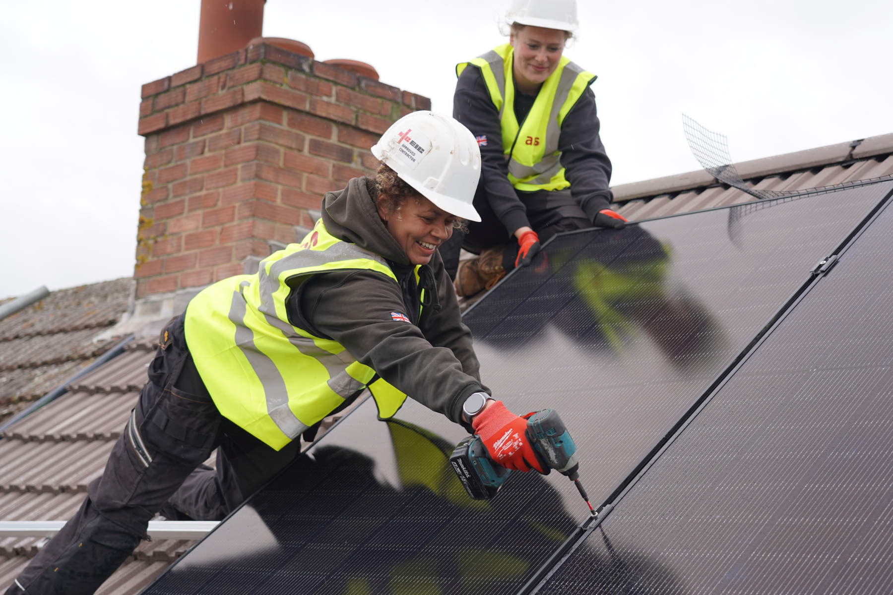 Two women in high viz jackets and hard hats installing solar panels on roof with tiles and chimney