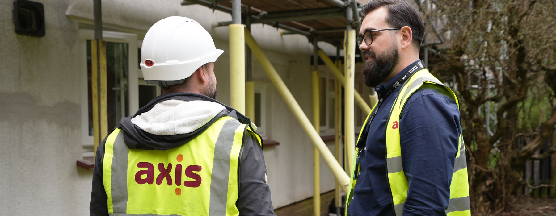Two men, both wearing yellow jackets with Axis logo. White helmet with a man turned to his back, the other with a beard and glasses facing side ways. Outside looking at scaffolding works