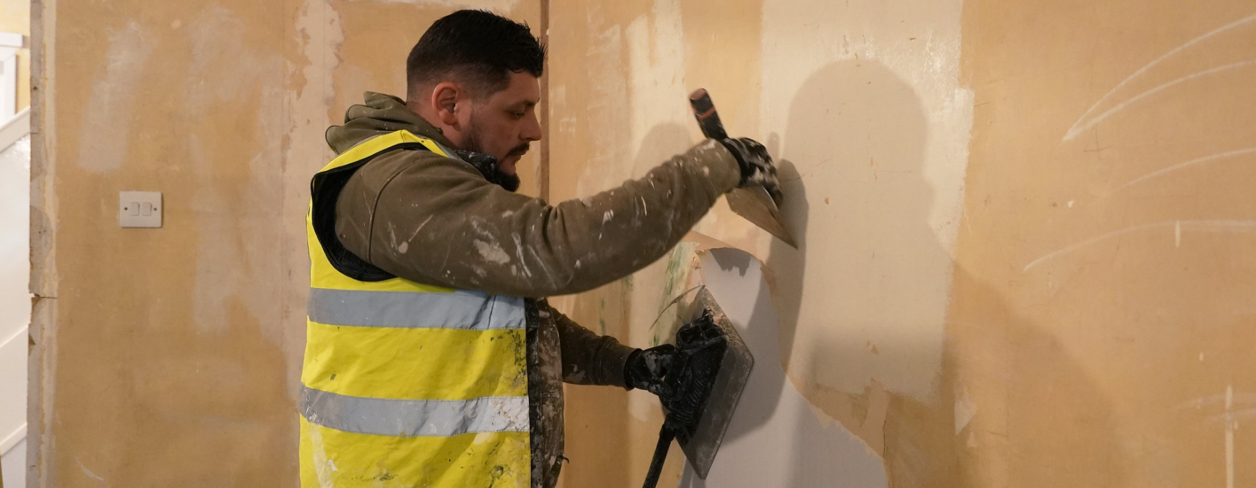 Multi operative man with a yellow vest and grey top with gloves refurbishing a wall