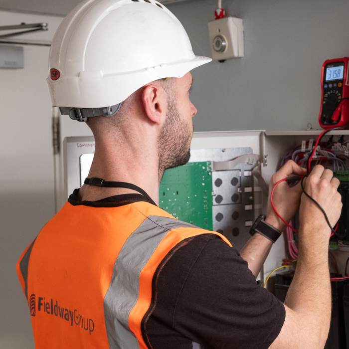 Man with white helmet, and orange vest with fieldway logo. Working with his hands to connect wires together on a panel on a wall