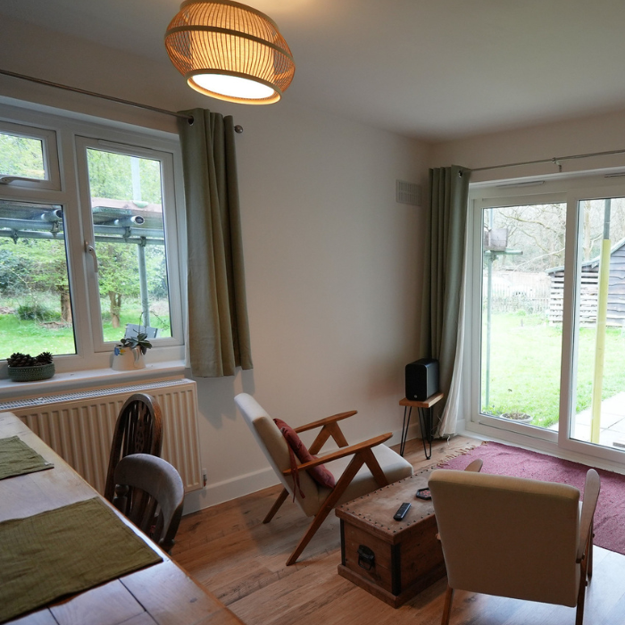 Inside a lodge sitting room. Brown chairs looking over of a large door with windows. Wooden table behind. Brown light fixture on the ceiling. Another window on the other wall. White walls. Wooden floor