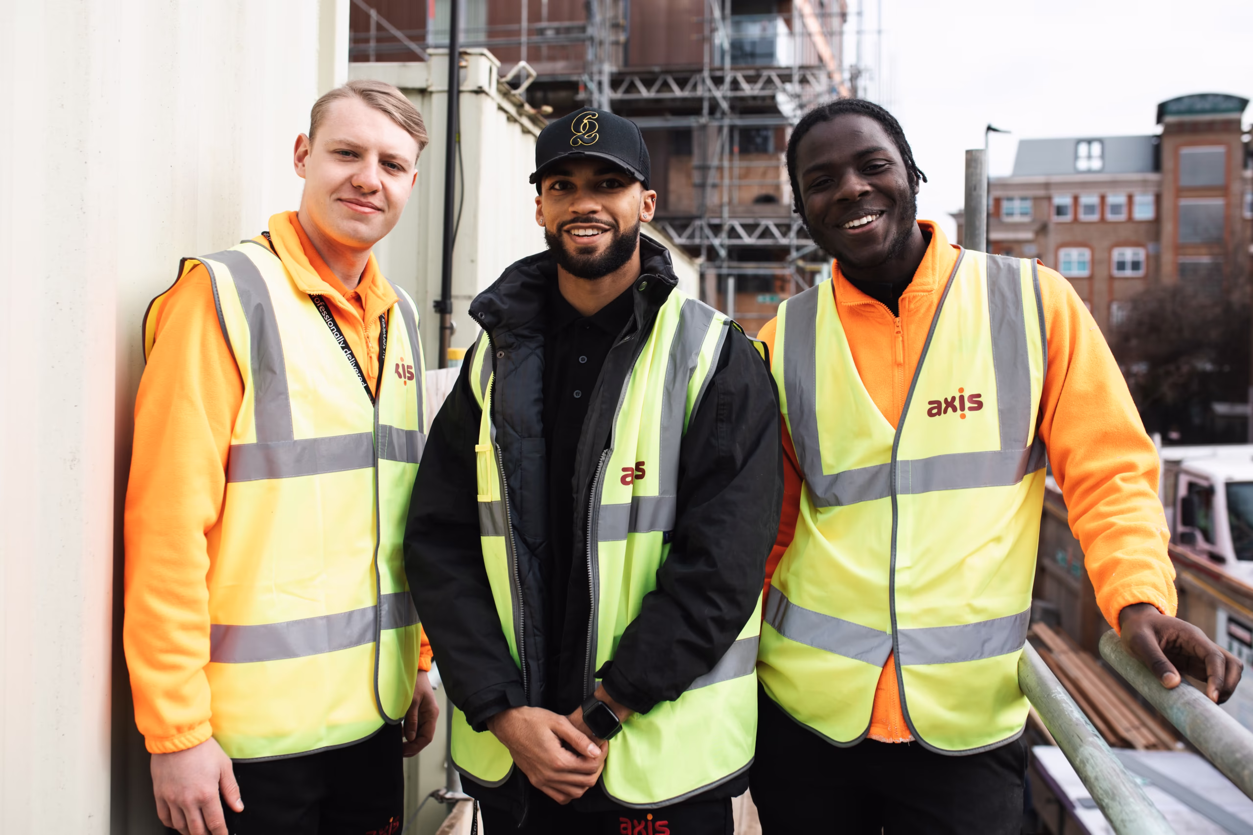 Three smiling Axis employees on a construction site surrounded by scaffolding.
