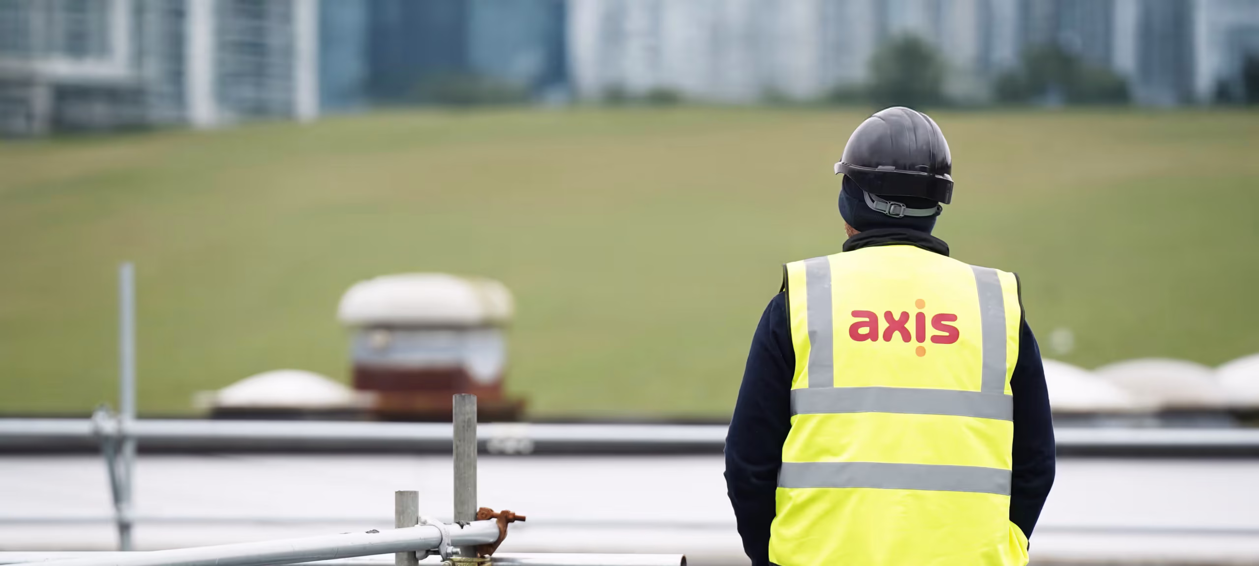 An Axis site worker in a hard hat and branded high-visibility vest, viewed from behind, surveys a rooftop work area with a cityscape in the background, representing information about Axis.