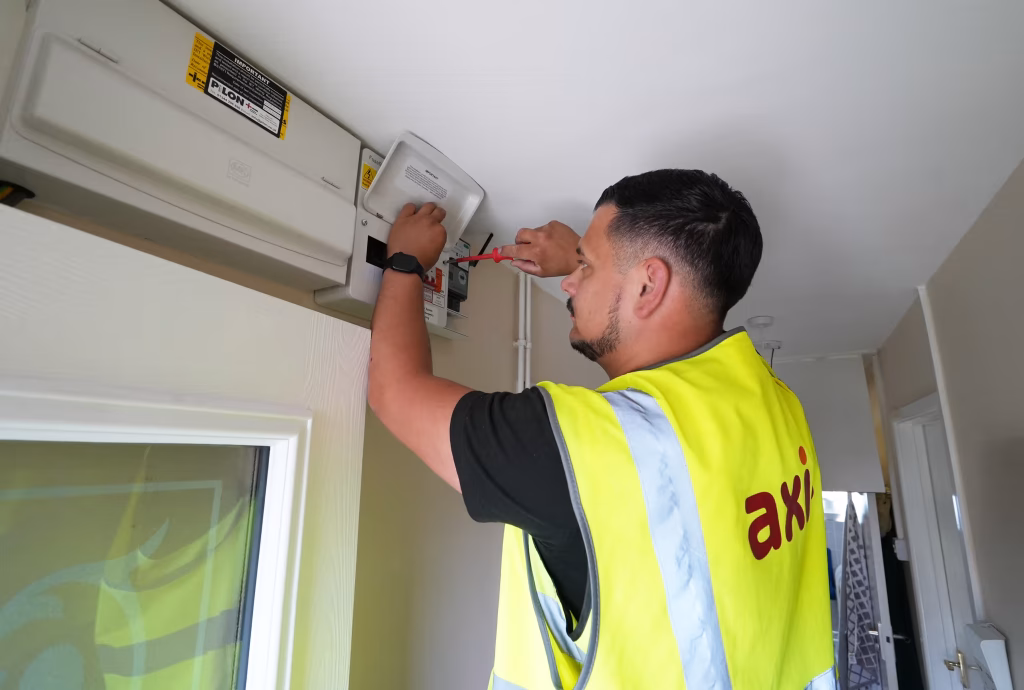 An Axis technician in a yellow high-visibility vest performs a repair on a wall-mounted electrical unit in a commercial building.