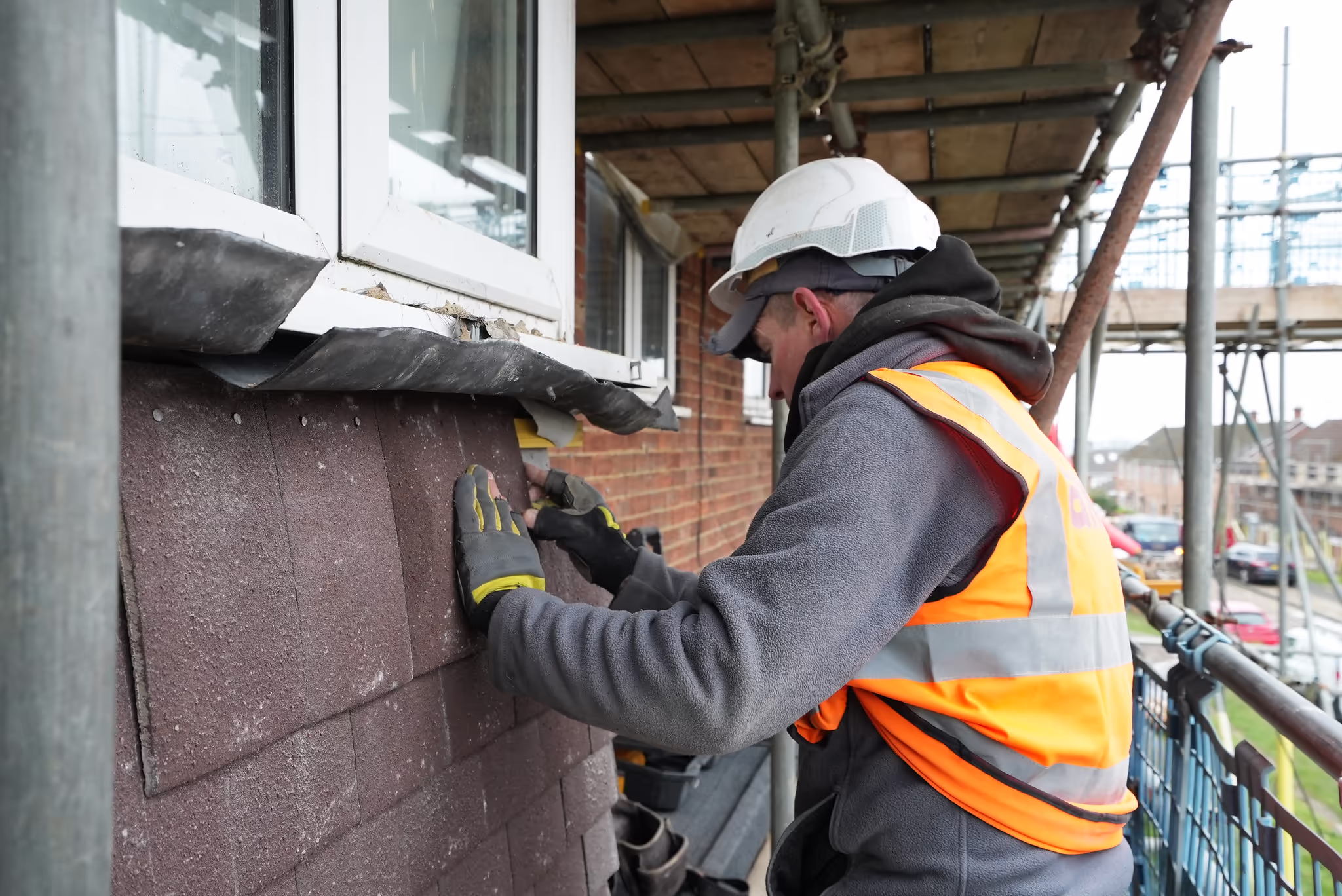 A skilled operative in a hard hat and safety gear works on scaffolding, carefully fitting tiles or cladding to the exterior of a house.