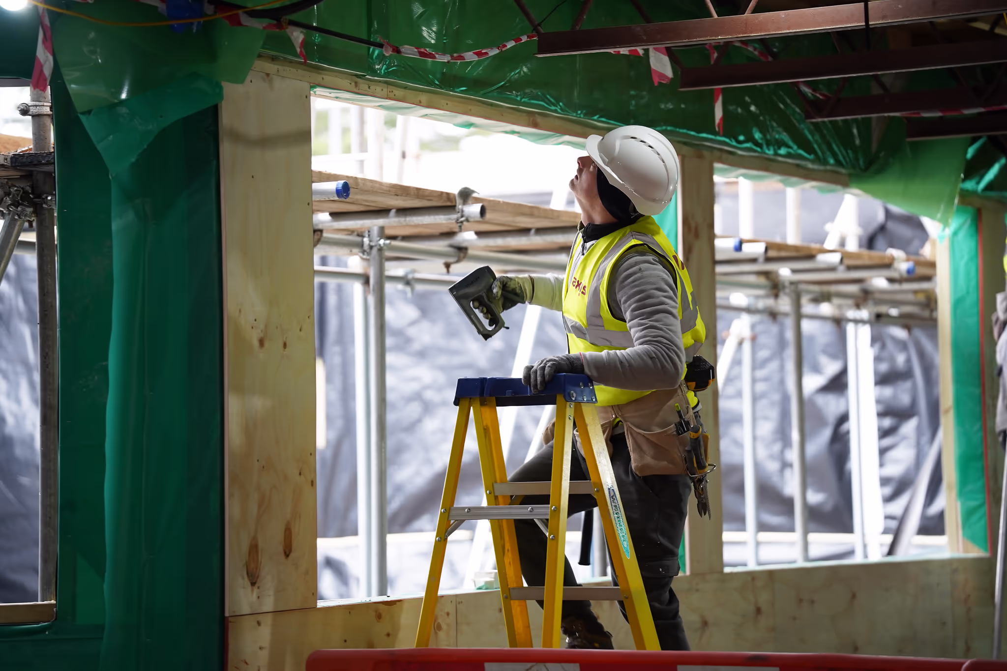 A construction worker wearing a hard hat and safety vest standing on a ladder and working near a window opening on a construction site.