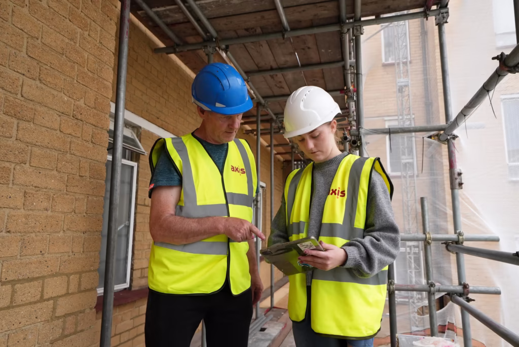 Two construction workers, one older with a blue hard hat and one younger with a white hard hat, reviewing documents on a construction site with scaffolding.