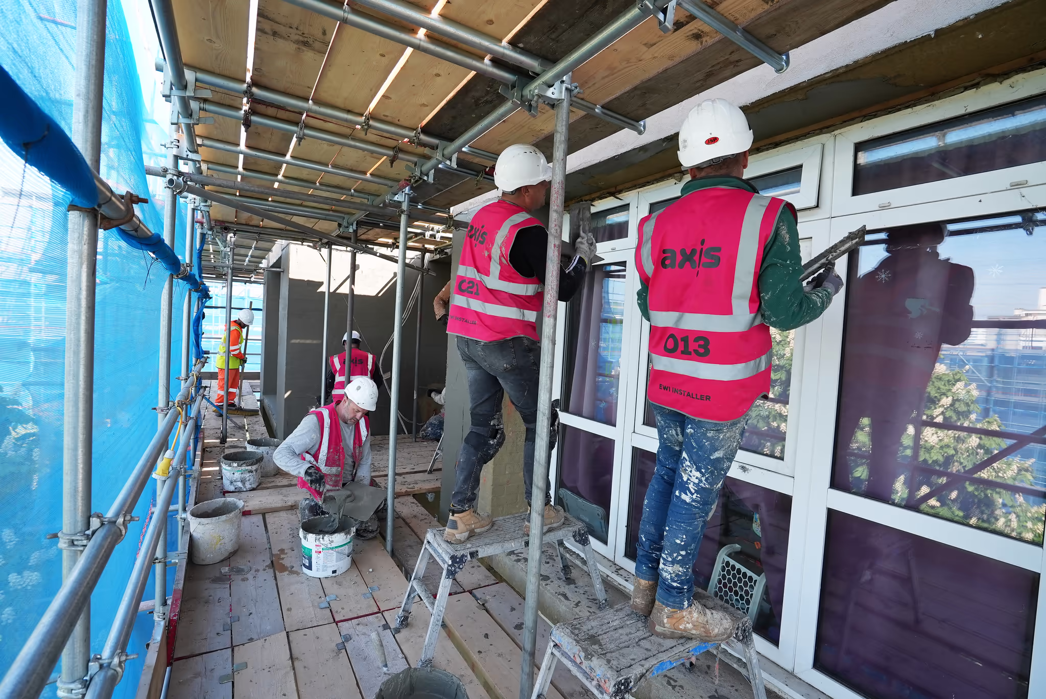 Construction workers in high-visibility vests and hard hats applying render or plaster to a building exterior from scaffolding.