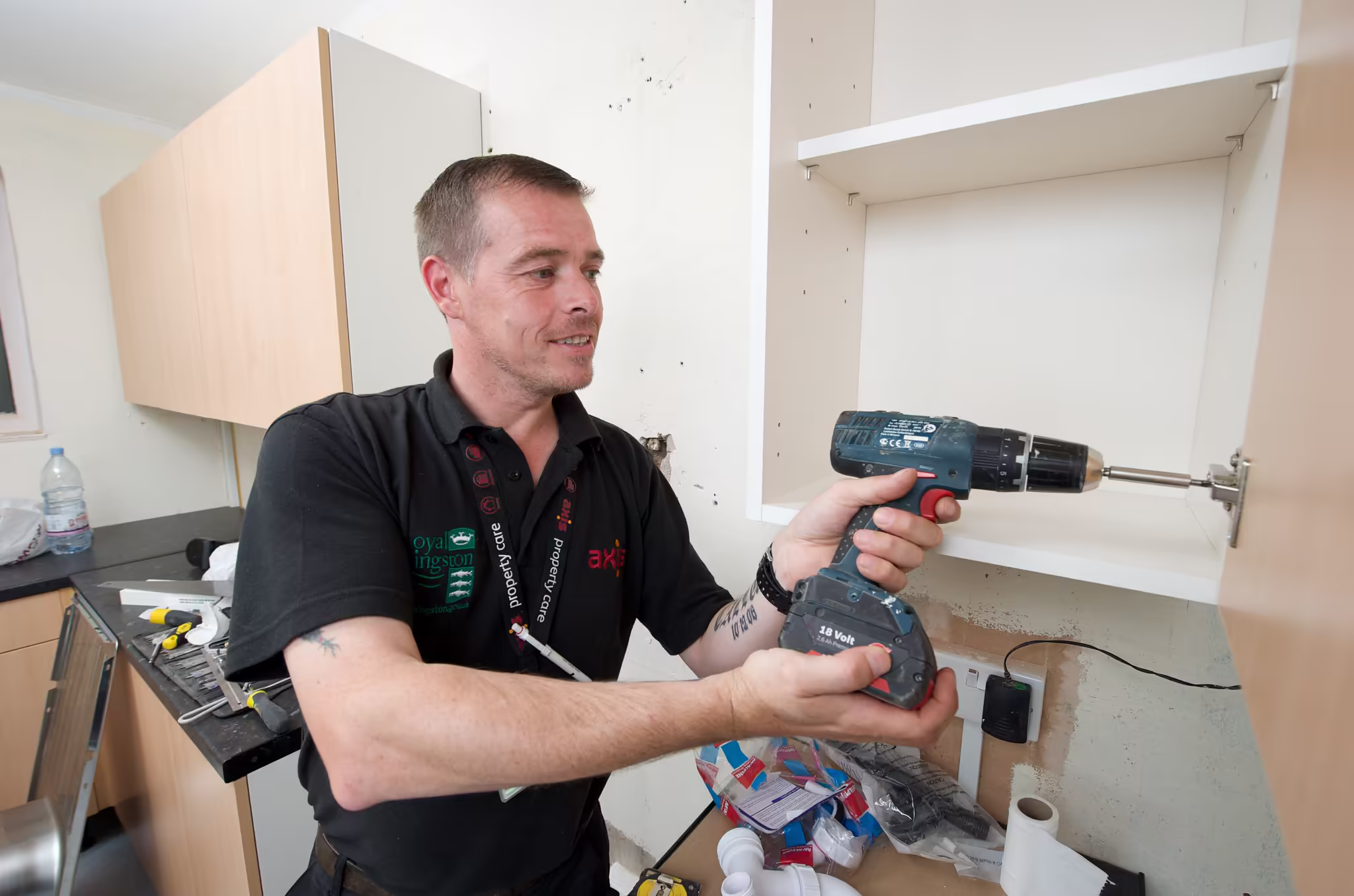 A smiling maintenance worker using a cordless drill to install a hinge on a kitchen cabinet in a home.