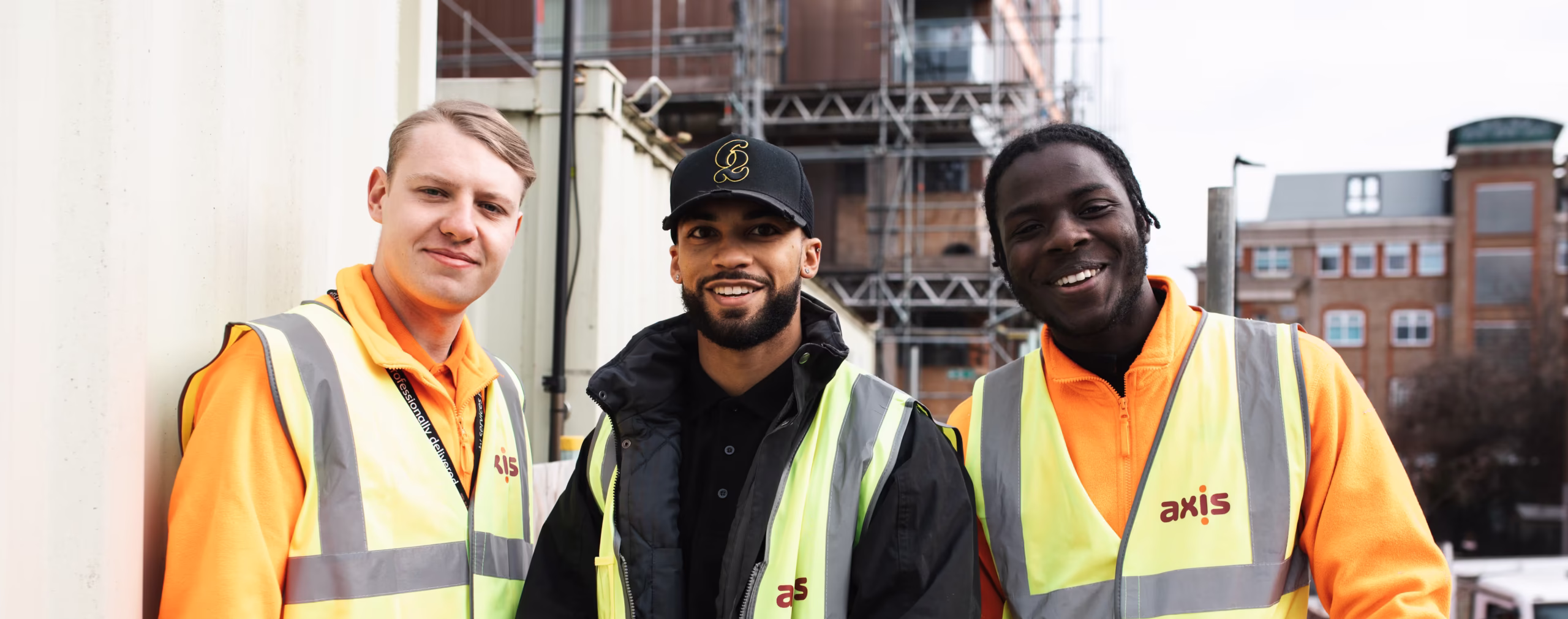 Three smiling Axis employees wearing high-vis vests on a construction site with scaffolding in the background.