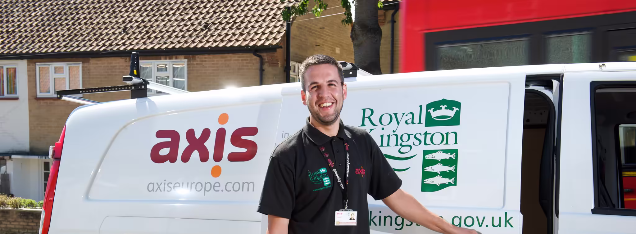 A smiling male tradesman wearing a black uniform shirt, standing by a white service van with a tool bag in his hand, representing subcontractors.
