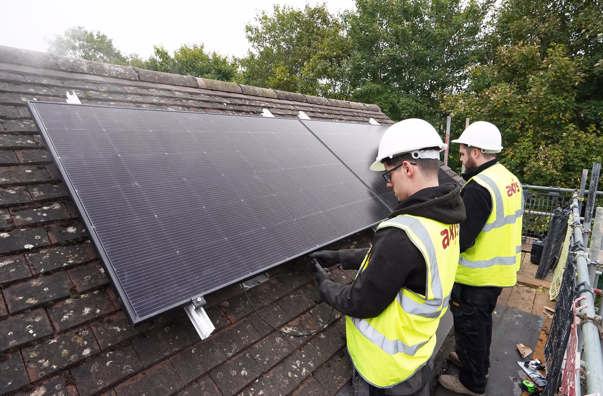Two workers in hard hats and safety vests installing black solar panels onto a tiled residential roof.
