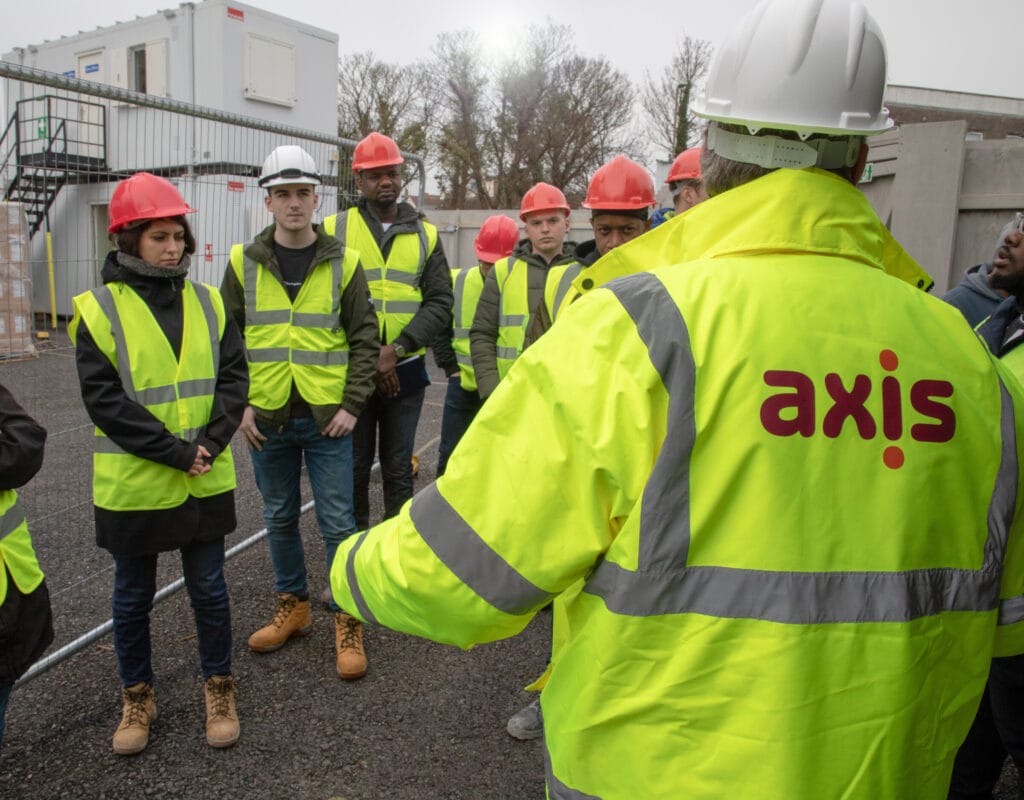 An Axis operative in high vis talking to eight young operatives on site surrounded by metal fencing.